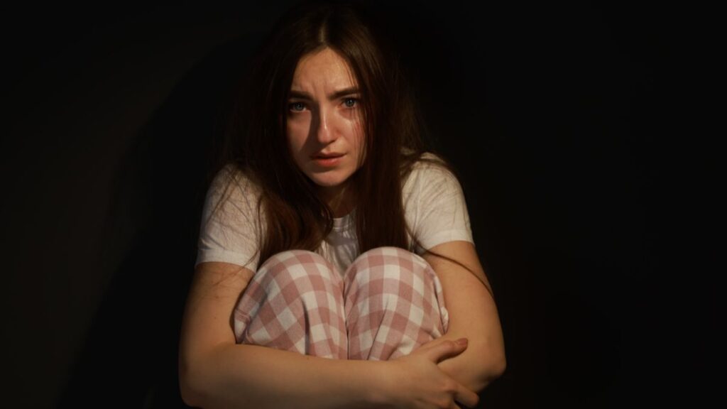 Frau sitzt ängstlich im Dunkeln, umarmt ihre Knie und blickt traurig nach vorn – Symbolbild für Sorgen und Ängste in der Trauer.